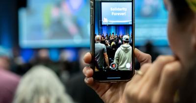 A delegate looks at their iPhone camera, which is displaying a stage where people are singing Solidarity Forever!